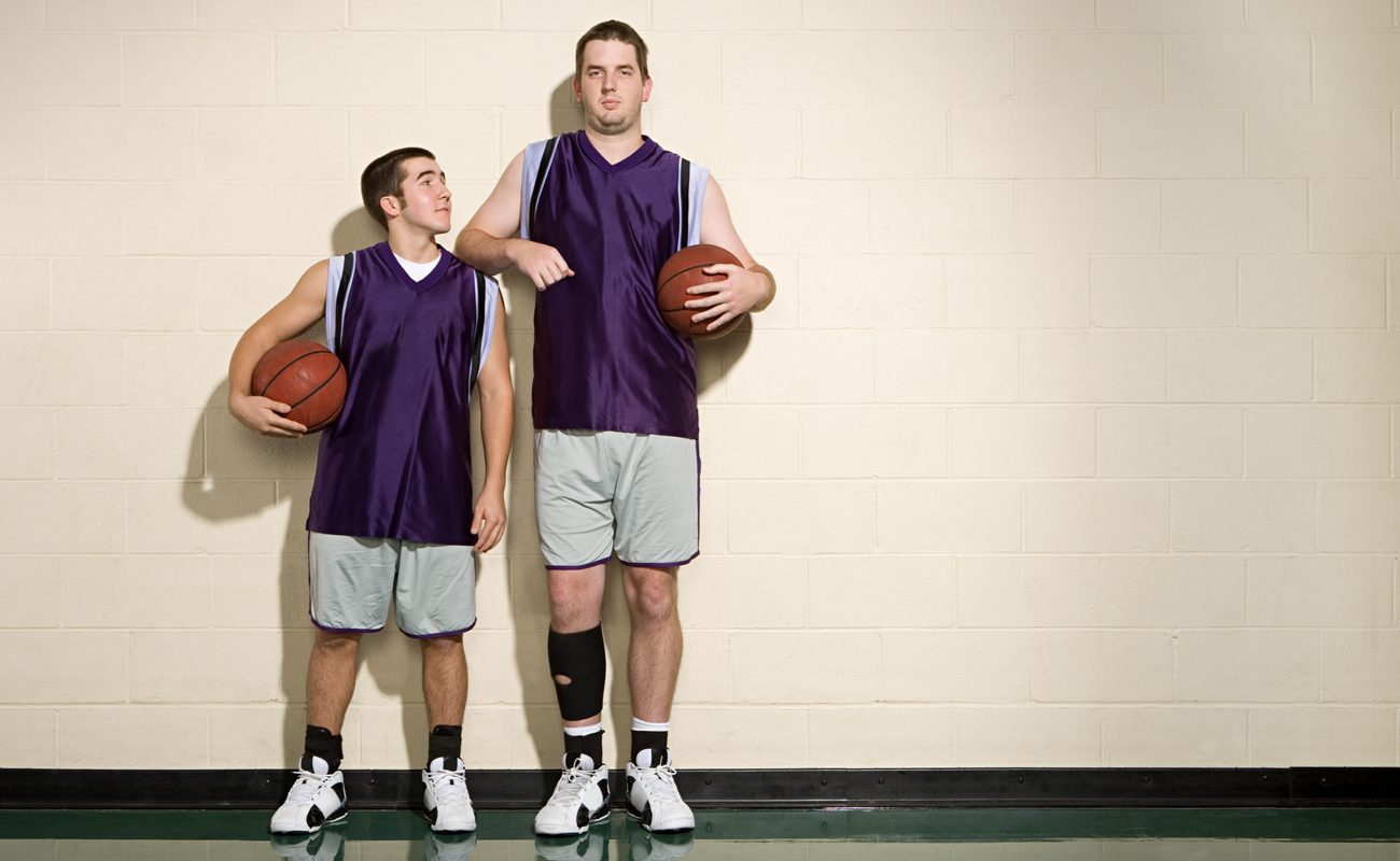 One short and one tall basketball players standing next to each other against a wall, both holding basketballs.